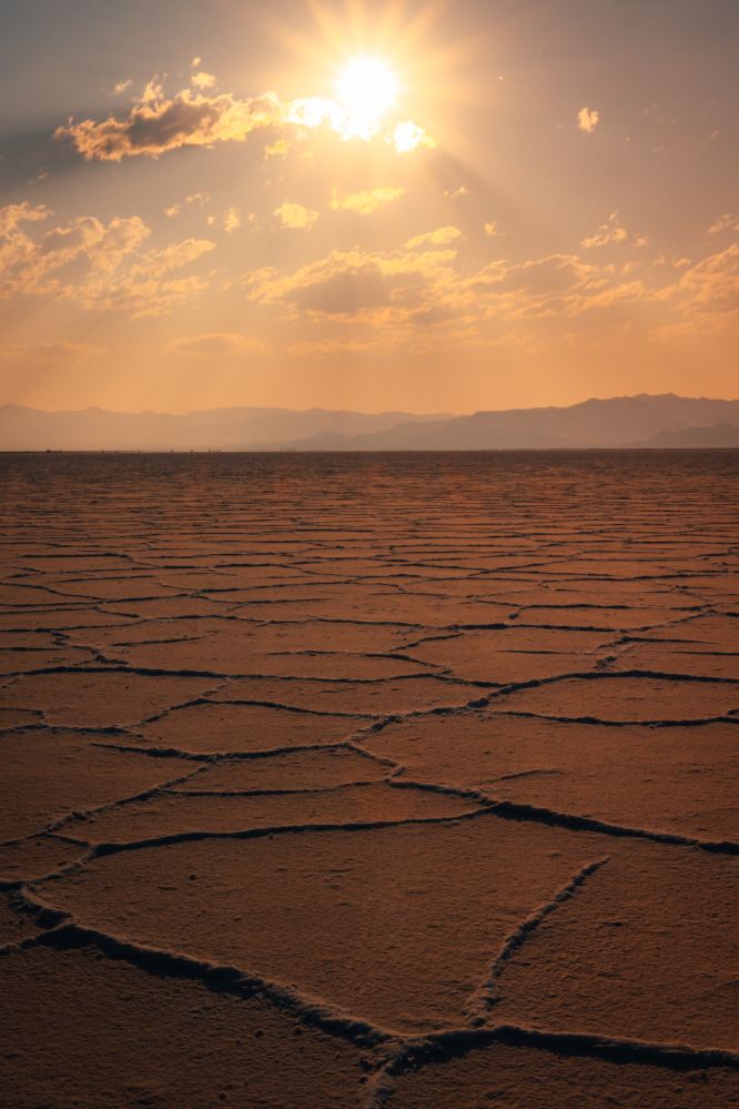 A desert. The ground is cracked. The sun is high with only a few clouds around it.