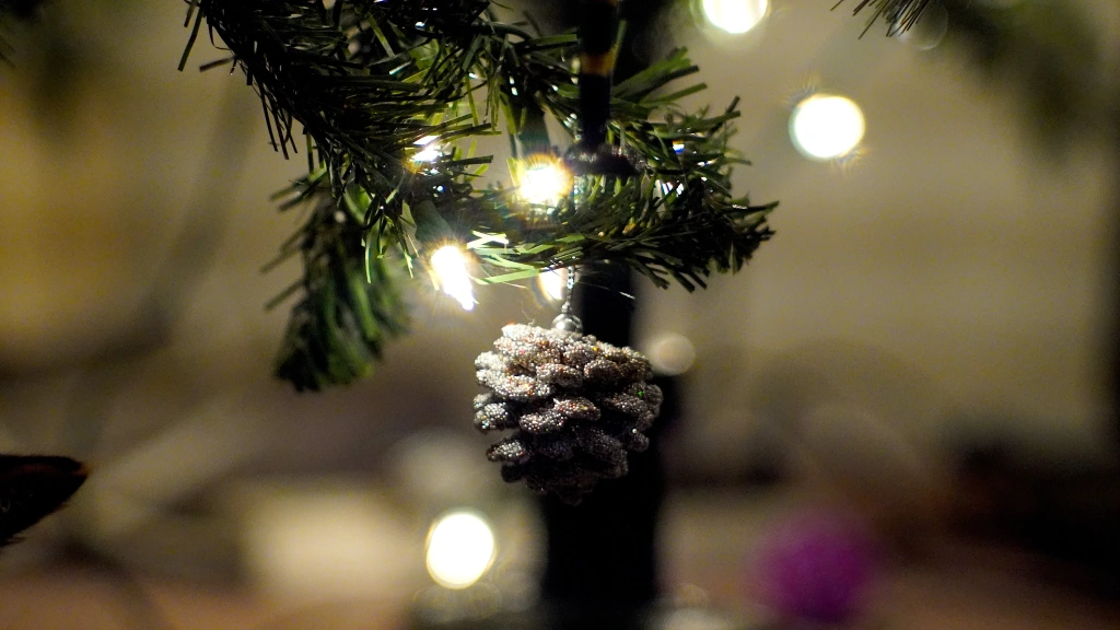 A close up of a sparkling pine cone ornament hanging from a softly lit Christmas tree to show warmth. 
